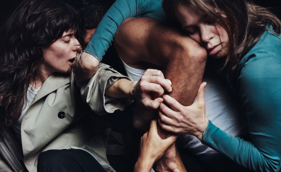 Fyra vuxna kvinnor ca 20–40 håller och böjer en mans ben och kropp i tät dansformation på mörk bakgrund.