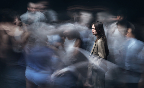 Kvinna ca 20–30 med långt brunt hår och beige kappa står stilla medan suddiga personer rör sig runt henne.