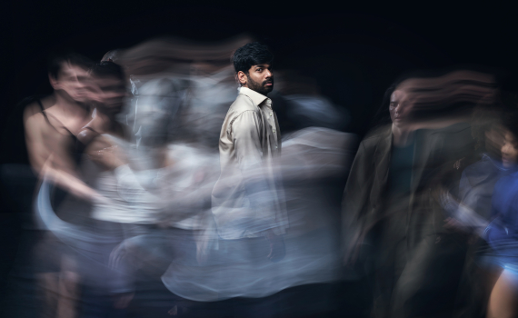 Man ca 25–35 med mörkt hår och beige skjorta står stilla medan suddiga personer rör sig runt honom.