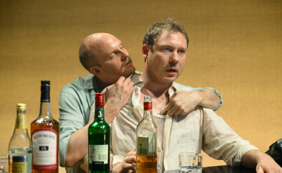 Två män sitter tätt vid ett bord med flaskor. Den ena håller den andra i skjortan i en laddad, intim scen.