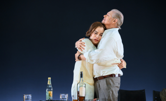 Man och kvinna omfamnar varandra nära ett bord med flaskor och glas. Scenen är stillsam och känsloladdad.