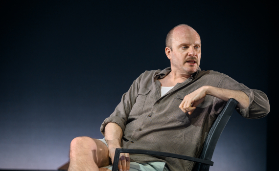 Man sitter avslappnat i en stol och ser åt sidan. Enkel scenbild med mörk bakgrund och få rekvisita.