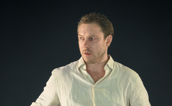 Man i vit skjorta står vid ett bord med flaskor och glas. Han ser åt sidan i en spänd scen.