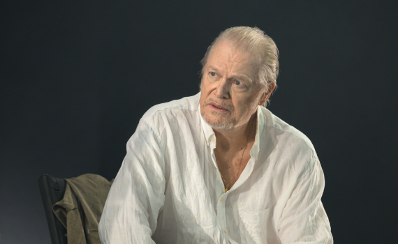 Äldre man sitter med ett glas i handen och ser upp. Porträttlik scenbild med dämpat ljus.