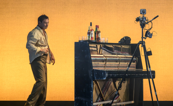 Man går förbi ett uppställt piano med flaskor ovanpå. En kamera på stativ står bredvid i en avskalad scen.