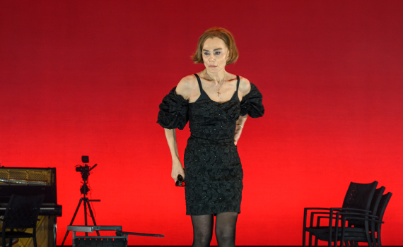 Kvinna i svart klänning går fram mot publiken mot en starkt röd bakgrund. Stolar och rekvisita står bakom.