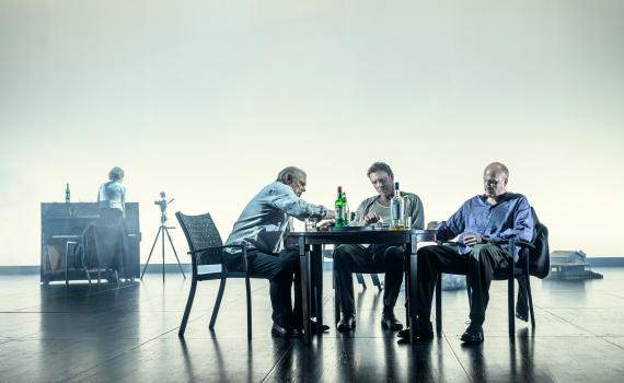 Tre män sitter vid ett bord med flaskor i ett öppet scenrum. I bakgrunden syns ett piano och en person vid sidan.
