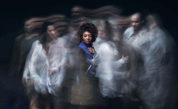 Svart kvinna ca 25–35 med afro och blå jacka står stilla medan suddiga personer rör sig runt henne.