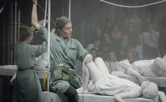 Kvinna ca 25–35 år i grön arbetsdräkt håller i trådar fästa vid ett barn målat vitt som ligger på ett bord, publik i bakgrunden.