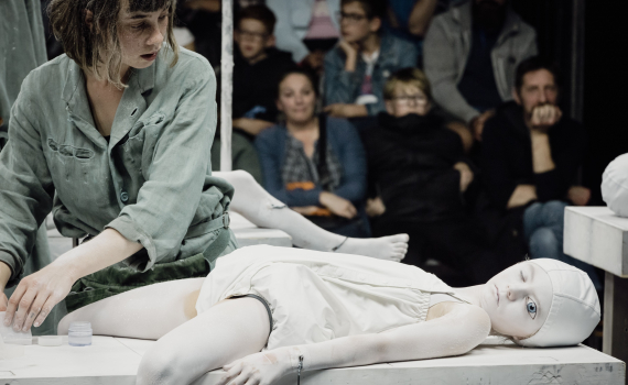 Kvinna ca 25–35 år i grön arbetsdräkt arbetar vid ett bord där ett barn målat vitt ligger på rygg, publik sitter bakom.