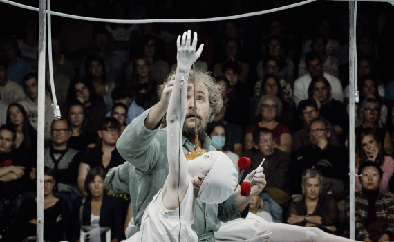 Man ca 40–50 år med lockigt hår och skägg i grön arbetsdräkt håller upp armen på ett barn målat vitt inför publik.