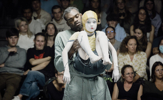 Man ca 30–40 år i grön arbetsdräkt bär ett barn målat vitt med gul hjälmmössa och randig tröja, publik bakom.