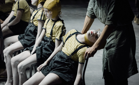 Flera barn målade vita sitter på stolar i gula hjälmmössor och randiga tröjor medan en vuxen i grön arbetsdräkt står bredvid.