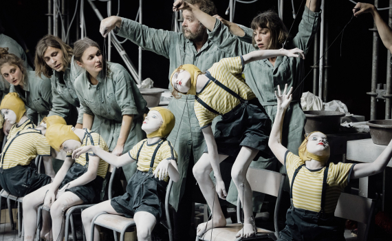 Fyra vuxna i gröna arbetsdräkter styr barn målade vita med trådar som marionetter på stolar, scenutrustning bakom.