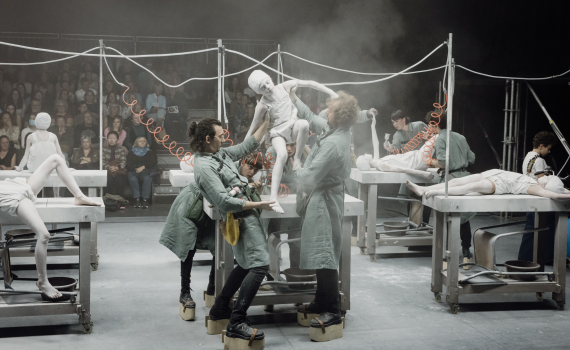 Scenföreställning där tre vuxna personer i gröna arbetskläder lyfter ett barn målat vitt på ett bord, publik sitter bakom.