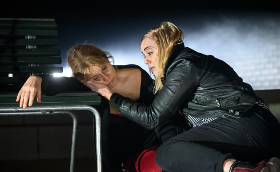 Två kvinnor ca 30–40 år sitter vid bord; blond kvinna i skinnjacka håller handen mot en annan kvinnas kind.