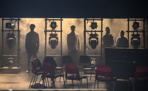 Fyra personer står i silhuett bakom scenlampor och rader av stolar på en teaterscen med rök.