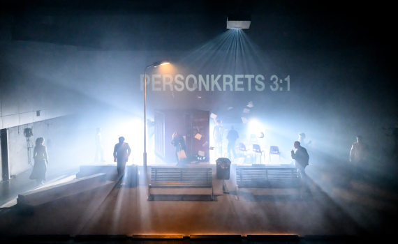 Scen med bänkar, gatlykta och container; flera personer rör sig i dimma, texten ”PERSONKRETS 3:1” projiceras.