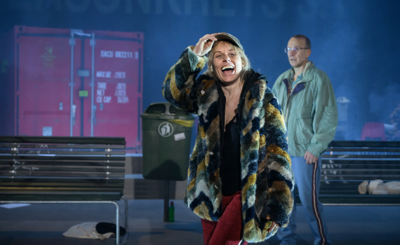 Kvinna ca 30–40 i keps, färgglad pälsjacka och röda byxor skrattar; man med glasögon står bakom.