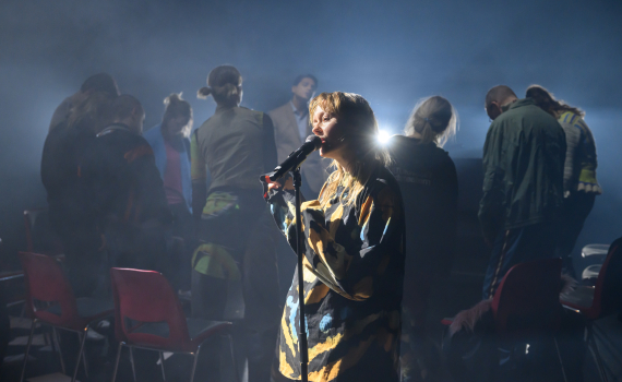 Kvinna ca 30–40 sjunger i mikrofon på scen; flera personer står bakom bland stolar i motljus.