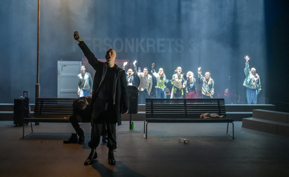 Man i svart rock står framför bänkar och håller upp armen; grupp människor bakom räcker upp händer.