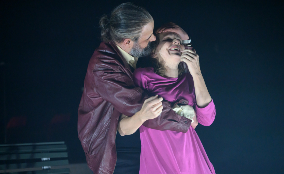 Man med grått hår i röd skinnjacka håller kvinna i rosa klänning bakifrån; de står tätt intill.