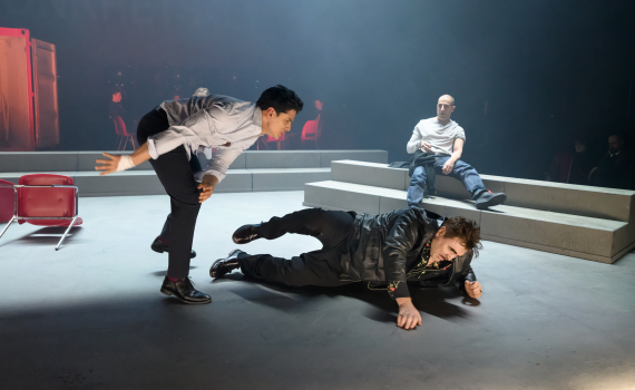 Man i vit skjorta sparkar man som ligger på golvet; tredje man sitter på trappsteg och ser på.