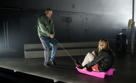 Man i glasögon drar kvinna i skinnjacka som sitter i liten rosa pulka med rep på scen.