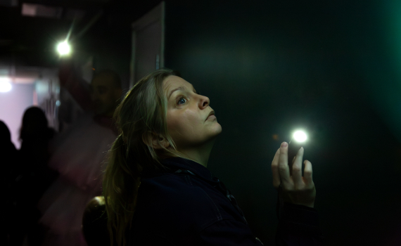 Skådespelare som lyser med en ficklampa i mörk korridor.