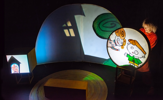 Scen med rund skärm där tecknade figurer projiceras; person i rött står bredvid.