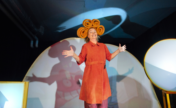 Person i röd klänning med spiralformad huvudbonad står med utsträckta armar framför rund scenbild.