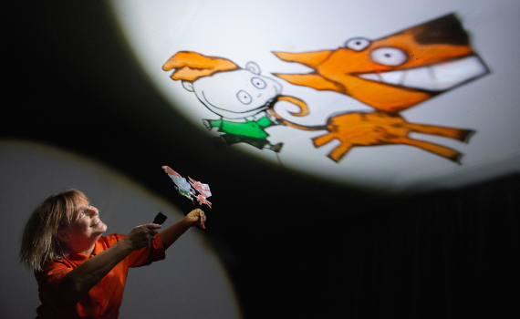 Person i röd klänning håller upp figurer som projiceras som tecknade bilder på en rund skärm i mörk scen.