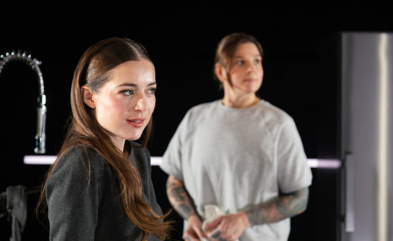 Två personer i ett mörkt rum; en kvinna i förgrunden ser åt sidan medan en person bakom henne står vid ett kök.