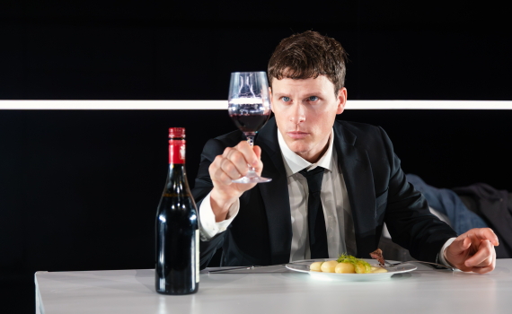 Man i kostym sitter vid bord och håller upp ett vinglas mot ljuset; en vinflaska och tallrik med mat står framför honom.