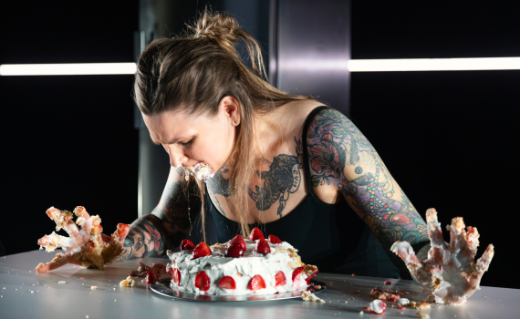 Tatuerad person lutar sig över ett bord och trycker ansiktet i en gräddtårta, med händerna nedkladdade i grädde.