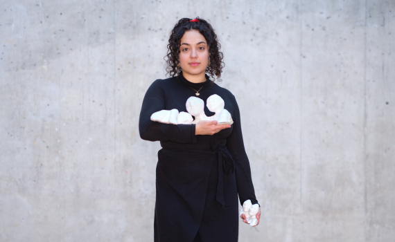 Person i svart klänning står mot betongvägg och håller vita, mjuka skulpturformer i famnen och handen.