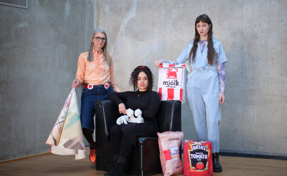 Tre personer poserar tillsammans; en sitter i fåtölj med vita objekt, två står med tygkassar och material runtom.