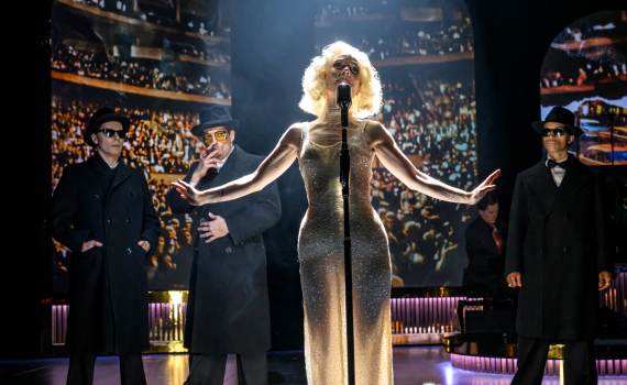 En kvinnlig sångare i glittrande lång klänning står i strålkastarljus vid en mikrofon på scen, med tre män i mörka rockar och hattar bakom sig och en publik projicerad i bakgrunden.