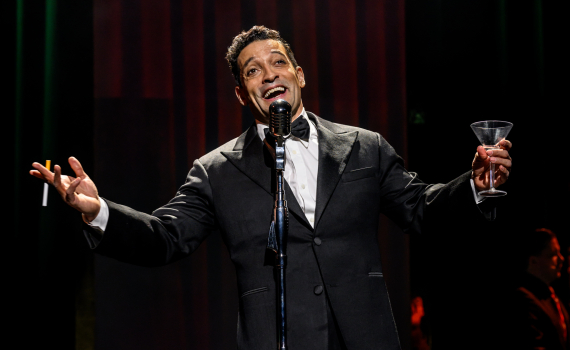 En man i smoking står vid en mikrofon och sjunger med öppna armar, håller en cigarett i ena handen och ett glas i den andra, mot en mörk scen med röd fond.