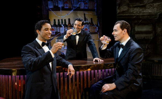 Tre män i smoking står vid en bar och skålar med whiskyglas, med flaskor uppställda bakom disken i en scenmiljö.