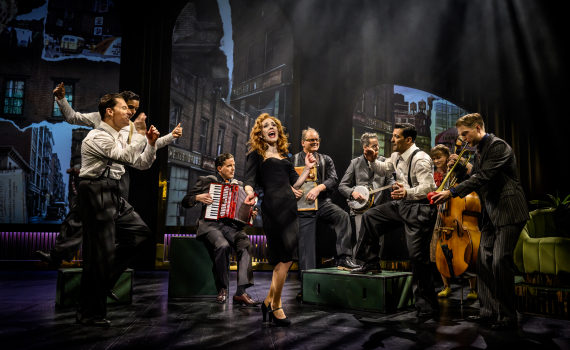 En ensemble på scen spelar och dansar; en kvinna i svart klänning sjunger i mitten medan musiker med dragspel, banjo och kontrabas omger henne i en stadsmiljö som fond.