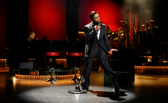 En man i smoking sjunger och rör sig dramatiskt vid en mikrofon, med ett band och röd bakgrund belyst bakom honom.