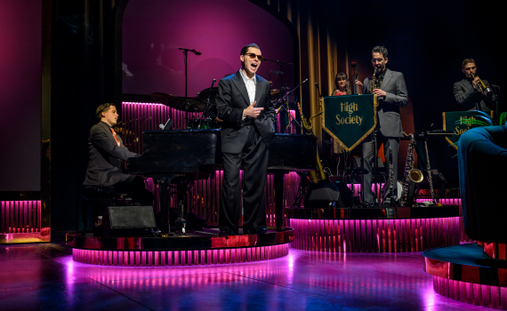 En sångare i smoking uppträder framför ett band på en scen med rosa belysning, där pianisten och blåssektionen syns bakom.