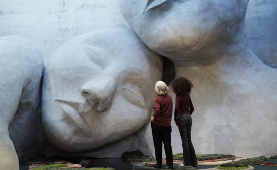 Två personer står framför en monumental skulptur med jättelika mänskliga ansikten i ett konsthallsliknande rum.