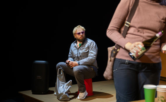 En man med lila solglasögon sitter på en pall medan en kvinna häller upp dryck i förgrunden.