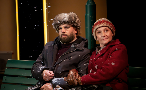 En man och en kvinna i vinterkläder sitter tätt tillsammans på en bänk medan snö faller omkring dem.