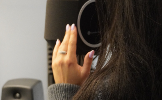 Person med långt mörkt hår håller handen mot en mikrofon i en ljudstudio. I bakgrunden syns högtalare och ljuddämpande paneler.