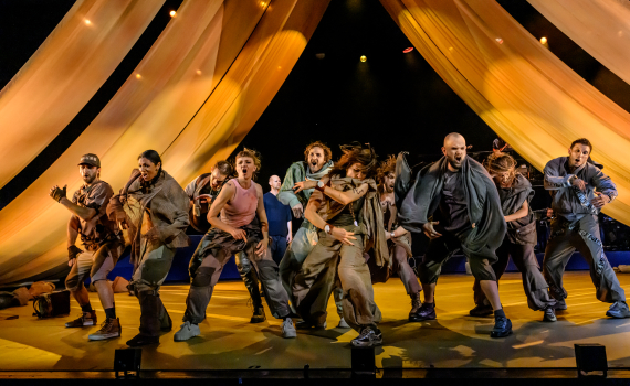 Ensemblen dansar tillsammans på scenen under stora guldfärgade draperier och varmt scenljus.