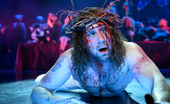 Närbild av Jesus med törnekrona liggande på scengolvet, med blod i ansiktet och ensemblen i bakgrunden.