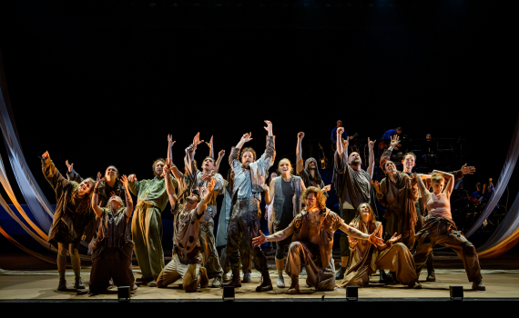 Ensemblen i Jesus Christ Superstar står samlad på scenen med armarna sträckta uppåt. Flera personer står på knä längst fram.